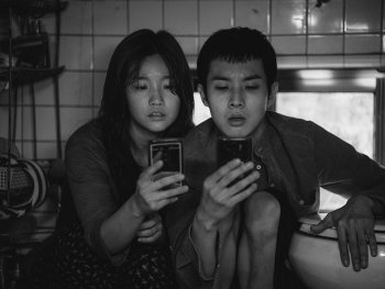 Copyright: Koch Films Eine junge Frau und ein Hunger Mann knien nebeneinander und schauen beide in ihre Handys. Das Foto ist schwarz-weiß.