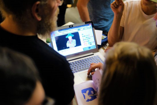 eine Gruppe von jungen Menschen steht um einen Laptop herum, auf dessen Bildschirm ein Spiel zu sehen ist.