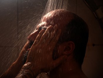 Ein Mann steht unter der Dusche und reibt sich mit den Händen im Gesicht.