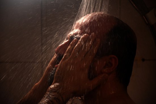 Ein Mann steht unter der Dusche und reibt sich mit den Händen im Gesicht.