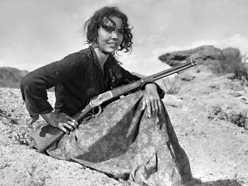 Schwarzweiß-Foto. Eine Frau sitzt mit einem Gewehr in der Hand auf einem Hügel.