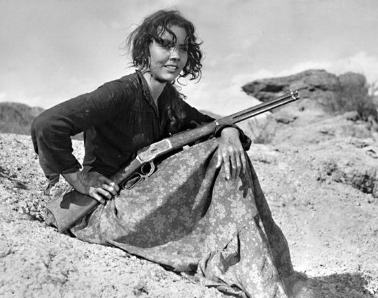 Schwarzweiß-Foto. Eine Frau sitzt mit einem Gewehr in der Hand auf einem Hügel.