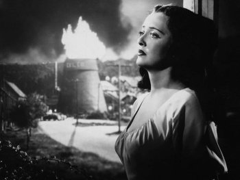 Schwarzweiß-Foto. Eine Frau lehnt an einem Fensterrahmen. Hinter brennt ein Turm.