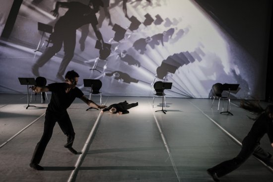 Weißer Bühnenraum, im Vordergrund Tänzer in Bewegung, im Hintergrund Tänzerin auf dem Boden liegend, vier Stühle, spiralförmig aufgefächterte Projektion des Bühnengeschehens auf Leinwand
