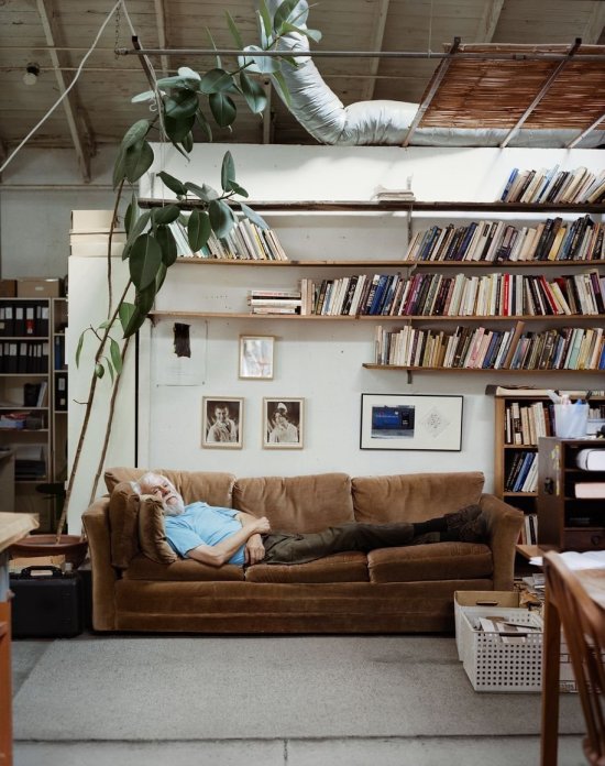 Älterer Mann liegt auf einem Sofa, das in einem Zimmer vor einer Wand mit Zimmerpflanze, Bildern und Bücherregalen steht