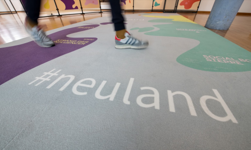 Person läuft in Turnschuhe über einen Teppich mit dem Schriftzug "#neuland".