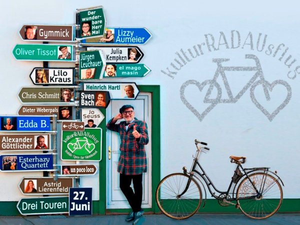 KulturRADausflug - beim Veranstalter Ein Mann steht neben einem Fahrrad und lehnt sich an ein Straßenschild mit vielen Künstlernamen