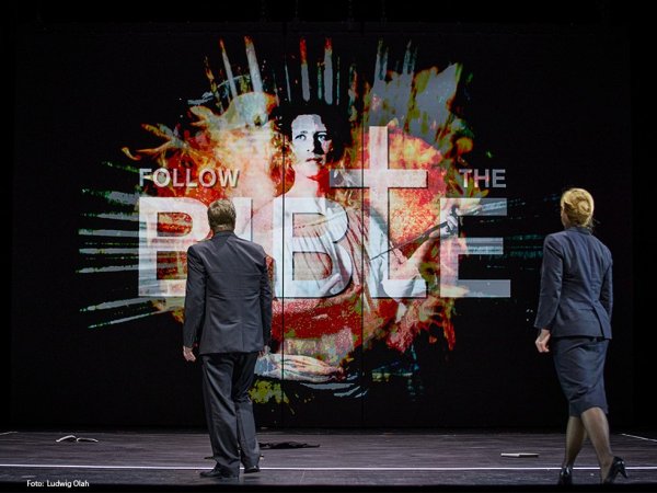 The Rape of Lucretia - beim Veranstalter Ein Mann und eine Frau betrachten eine Projektion mit dem Text "Follow the bible"