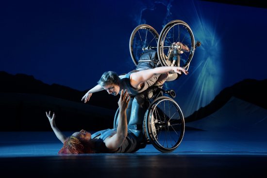 Die Tänzerin Laurel Lawson fliegt mit ihren ausgestreckten Armen durch die Luft. Sie wird von der Tänzerin Alice Sheppard gestützt, die sich von unten hochhebt. Sie schauen einander an und lächeln. Beide sitzen in einem Rollstuhl.