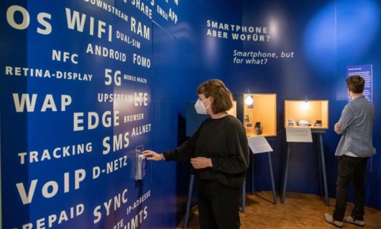 Blick in den neuen Ausstellungsbereich: Eine Frau mit Maske nimmt ein Infoblatt und ein Mann mit Maske betreachtet eine Vitrine. Die Vitrine steht vor einer blauen Wand.
