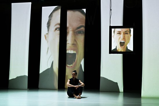 Dies ist ein Foto einer Frau, die auf der Bühne sitzt, sie scheint zu schreien und es gibt mehrere Projektionen hinter ihr von ihrem Schreien.