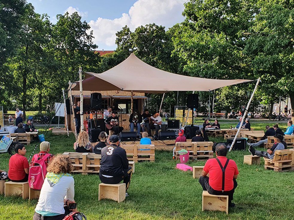 KommVorZone - KUF im südpunkt Bühne im Park mit Besucherinnen und Besuchern, welche auf Holzkisten davor sitzen