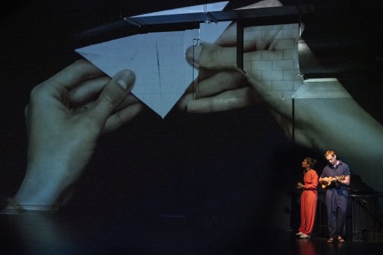 Zwei Schauspieler an der rechten Bildseite, im Hintergrund eine Hand, die Origami-Figuren faltet an die Wand gebeamt