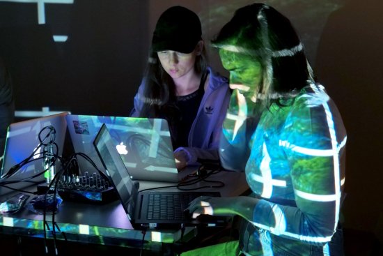 Zwei Frauen an einem Laptop überlagert von einer Lichtinstallation