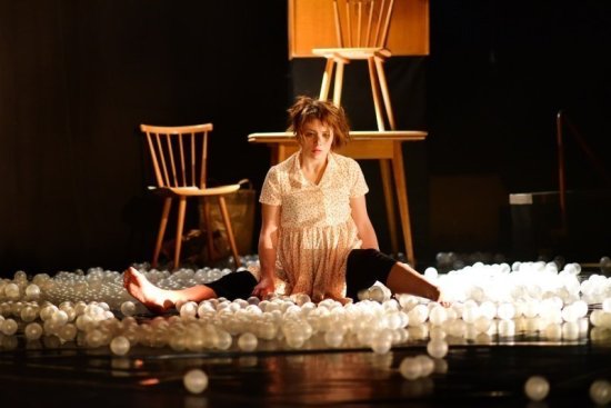 Frau sitzt auf einer Bühne umgeben von weißen Plastikbällen, im Hintergrund stehen Tisch und Stühle