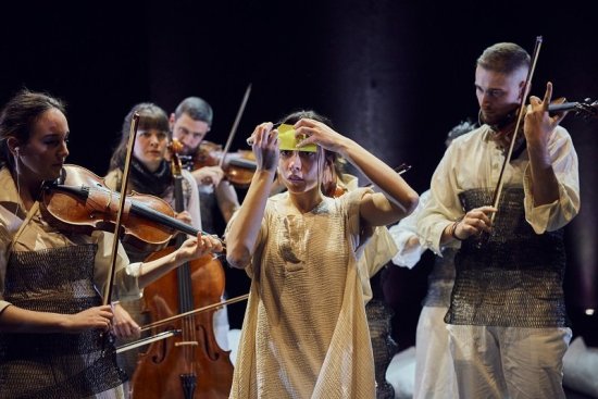 Eine junge Frau ist umringt von Musiker*innen mit ihren Instrumenten. Die Frau klebt sich einen gelben Klebezettel auf die Stirn.