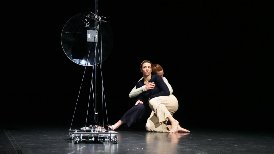 Zwei Tänzerinnen blicken eng umschlungen auf einen Roboter.