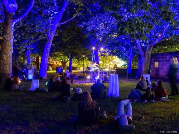 Zuhörerinnen und Zuhörer sitzen am Abend an der Stadtmauer auf einer bunt beleuchteten Wiese unter Bäumen und hören einem DJ zu.