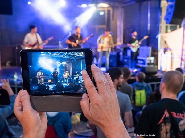 Jemand filmt mit einem Smartphone eine Musikband auf einer Bühne.