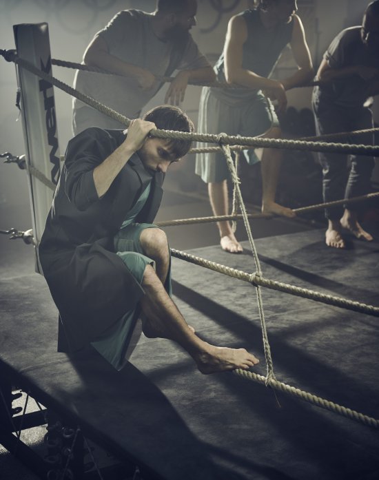 Tänzer hockt auf der Umrandung eines Boxrings aus quer gespannten Seilen, hält sich mit einer Hand fest, schummriges Licht; im Hintergrund lehnen sich mehrere Personen über die Begrenzung des Boxrings