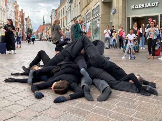 Mehrere Menschen in schwarzer Kleidung liegen in einer belebten Fußgängerzone als Haufen übereinander