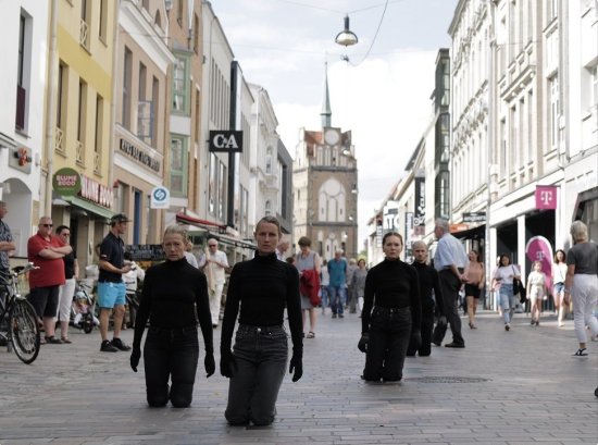 Vier Performende in schwarzer Kleidung knien in einer belebten Fußgängerzone, Blick in die Kamera