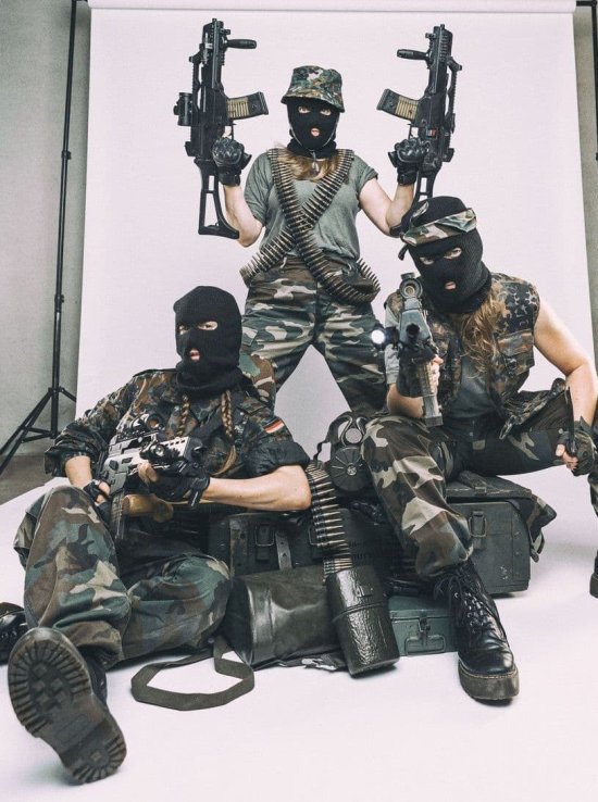 Drei Personen in Militärkleidung mit Sturmmasken und Maschinengewehren vor einem Foto-Rollup, Person in der Mitte steht, eine hockt, eine liegt
