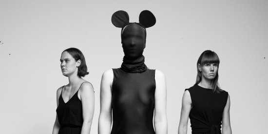 schwarz-weiß Bild, mittig eine Person mit schwarzer Maske, die das ganze Gesicht bedeckt und Mickey Mouse Ohren, links und rechts die Tänzerinnen Eva Borrmann und Evelyn Hornberg, die zur Seite schauen