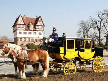 Zwei Pferde. Eine gelbe Kutsche mit einem Mann in blauer Uniform vor einem Schloss in rotem Fachwerk. Dahinter Bäume ohne Laub