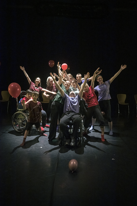 Zu sehen sind Tänzerinnen und Tänzer des Amateur-Ensembles von EveryBody. Sie stehen gut gelaunt zusammen auf einer Bühne, recken lachend die Arme in die Luft und posieren. Manche von ihnen haben eine sichtbare Behinderung und benutzen zum Beispiel einen Rollstuhl. Um sie herum sind Luftballons.