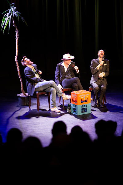 Jan Bratenstein, Marti Mäkkelä und Tim Steinheimer auf der Bühne, lachend mit Mikros, im Hintergrund eine Palme, vor ihnen stehen zwei Getränkekästen als Tisch