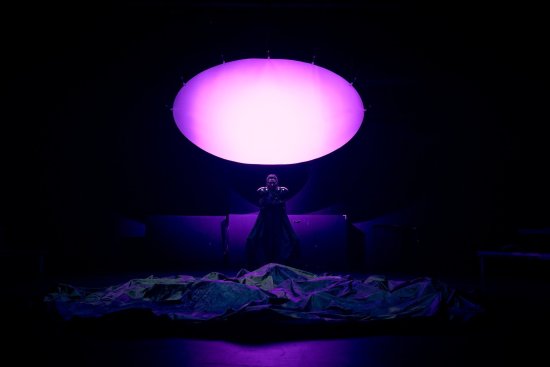 eine Performerin als Cyberwitch, vor ihr ein großes faltiges dunkles Tuch, über ihr ist eine mit violettem Licht angestrahlte Projektionsfläche, der Raum ist sehr dunkel