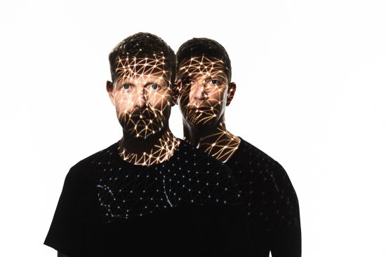 Portraitfoto von Nils Wülker und Arne Jansen vor einem weißen Hintergrund, sie werden mit Lichtprojektionen angestrahlt