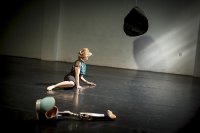Tänzerin mit einem Bein halbliegend auf dem schwarzen Tanzboden; ihre Prothese liegt nebendran