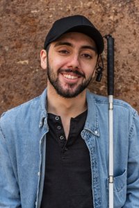 lächelnder Mann mit schwarzem Käppi und Jeanshemd, links von ihm ein Blindenstock