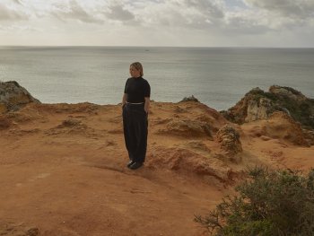 junge Frau mit schwarzen Hosen an der Küste auf Felsen, im Hntergrund das Meer