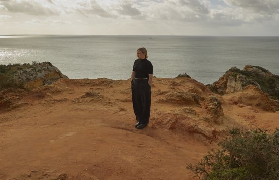 junge Frau mit schwarzen Hosen an der Küste auf Felsen, im Hntergrund das Meer
