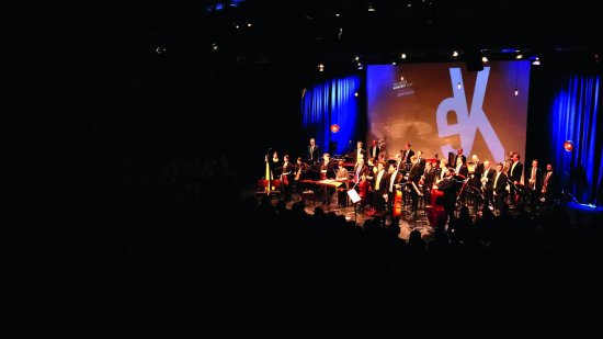 Orchester auf einer Bühne, vor blauem Vorhang, dahinter auf einer Leinwand die Buchstaben eK