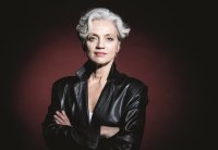 Frau mit weißem Kurzen Haar, schwarzer Lederjacke vor dunkelrotem Hintergrund. lächelnd