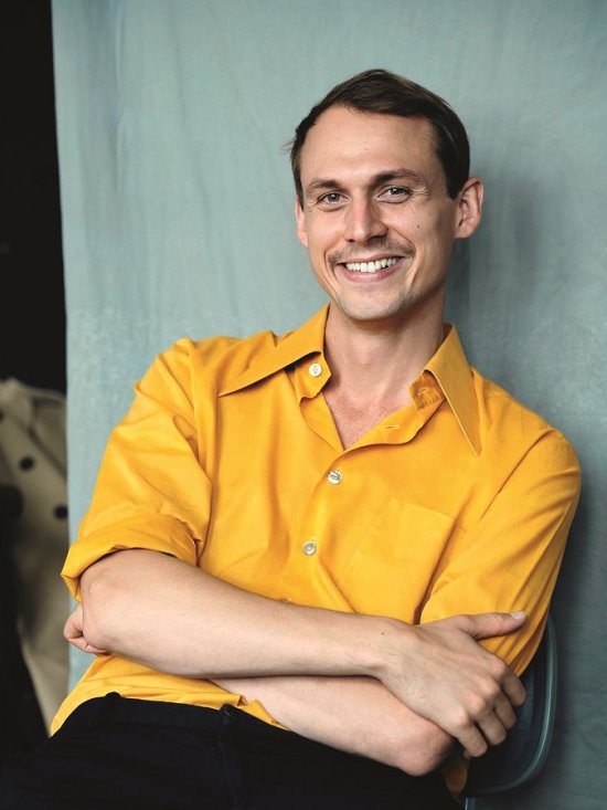 lächelnder junger Mann mit kurzem dunklen Haar, verschränkten Armen und gelbem Hemd