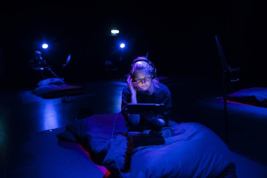 junge Frau sitzt im Dunkeln auf Futon mit Kophörern vor einem Bildschirm.