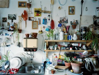 Blick auf eine Küchenwand mit Spüle und Kühlschrank im Stl der 1960er-Jahre,  in der alle Flächen mit Geschirr und Pflanzen zugestellt sind. Die Wand wird als Pinwand für Fotos und kleinen Erinnerungsstücke genutzt