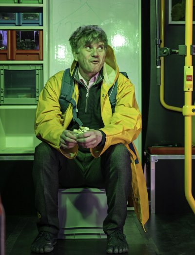 Mann, der eine gelbe Regenjacke und einen Rucksack trägt, sitzt auf einer umgedrehten Kiste auf der Spielfläche im Theaterbus