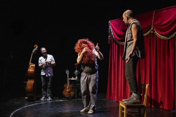 Drei Schauspieler vor einem roten Vorhang, einer auf einem Stuhl, einer mit roter Perücke und Bart