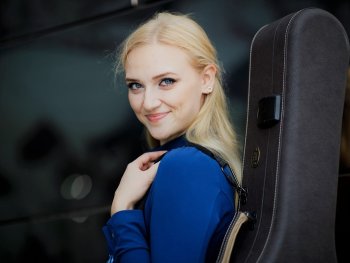 Eine blonde Frau mit stechend blauen Augen trägt einen Gitarrenkoffer. Sie schaut über ihre Schulter in die Kamera.