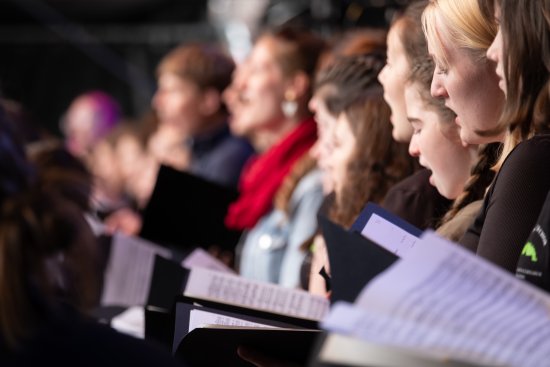 Mehrere Personen stehen in einer Reihe, alle halten Notenblätter in den Händen und scheinen zu singen.