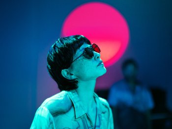 Eine Person im Hemd und mit Sonnenbrille schaut nach rechts oben und wird von einem blauen Licht angestrahlt. Im Hintergrund ist eine runde, rötliche Lichtquelle und eine unscharfe Person zu sehen.