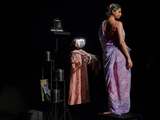 In der rechten Bildhälfte steht Mallika Taneja in einem langen lila Gewand mit dem Rücken zu uns und über ihre rechte Schulter blickend. Links Neben ihr steht eine etwas kleinere Puppe und daneben ein Metallgestell mit Büchsen darauf. Sie warden von einem Scheinwerfer von rechts angestrahlt, der Hinter- und Untergrund ist Schwarz.