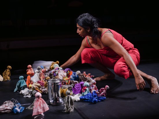 Mallika Taneja sitzt in der rechten Bildhälfte in der Hocke auf einer schwarzen Bühne und vor schwarzem Hintergrund. Sie beugt sich zu unzähligen bunten Stoffpuppen herunter, die in einem Haufen zusammenliegen oder dahinter auf dem Boden stehen.
