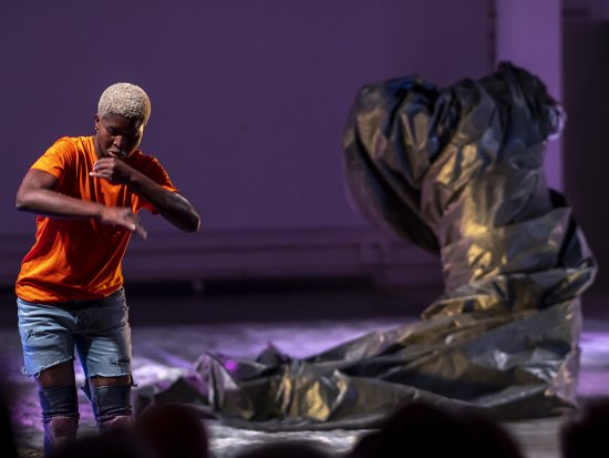 Eine Person tanzt in einem orangenen T-Shirt, einer Jeansshorts und Knieschonern gekleidet vor einer aufrechtstehenden, verschlungenen, schwarzen Plastikplane. Der Hintergrund ist lila-pink beleuchtet.